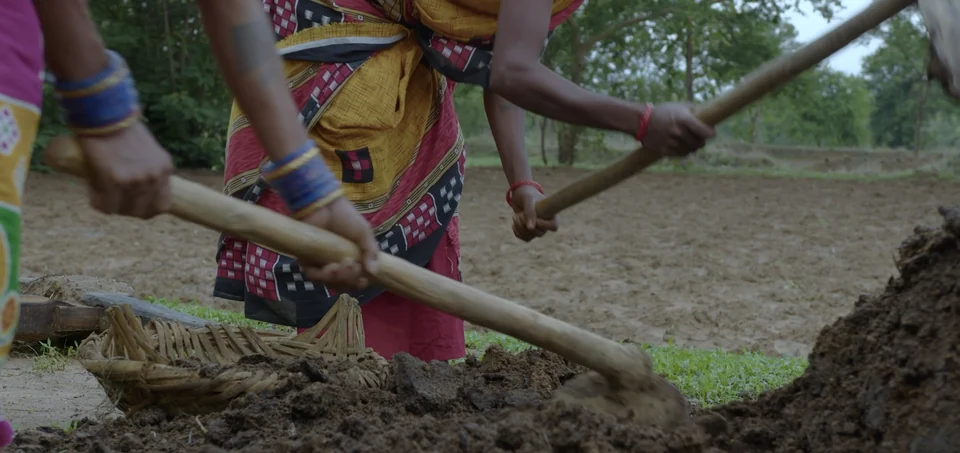 Hands working compost with wooden-handled tools beside a field
